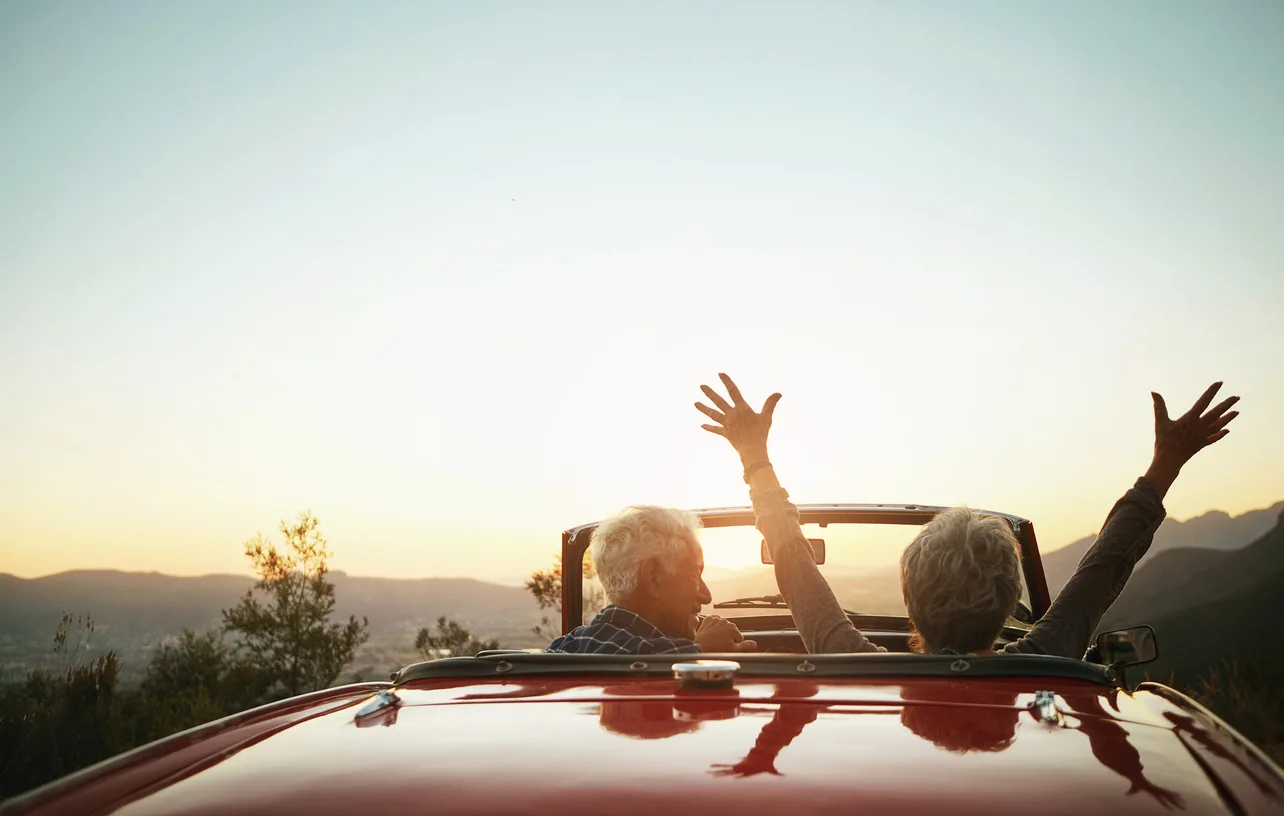 Two older adults riding in a red convertible at sunset